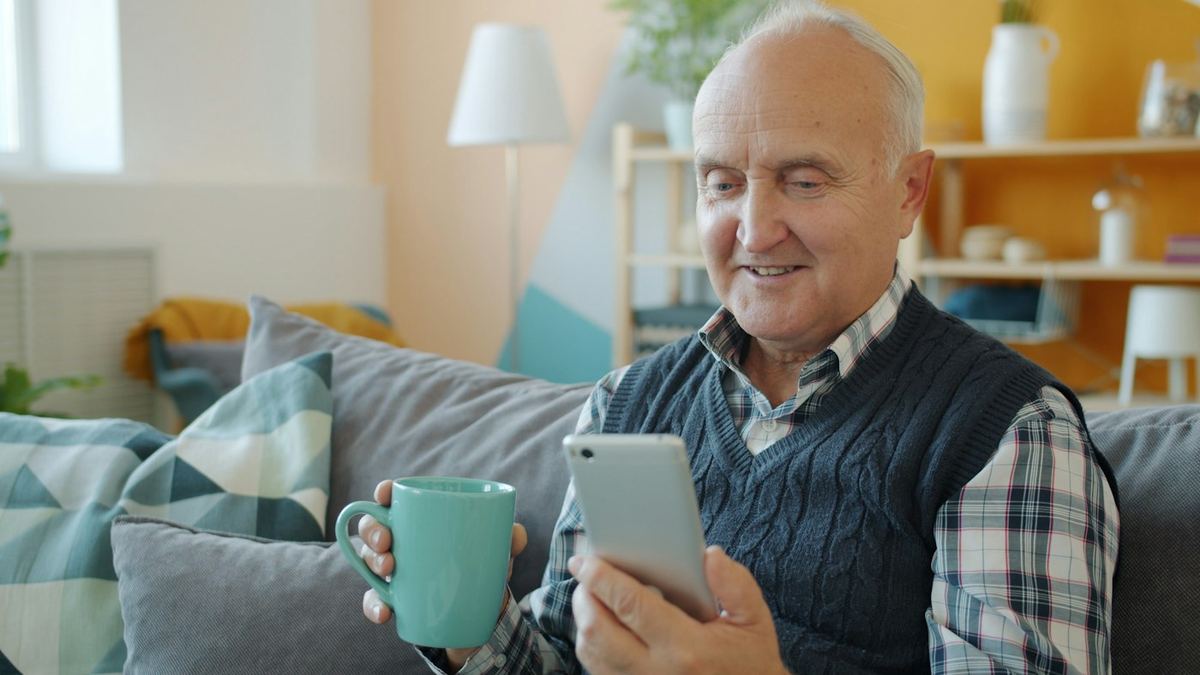 Homme âgé souriant sur un canapé, une tasse de thé dans une main et son téléphone dans l'autre
